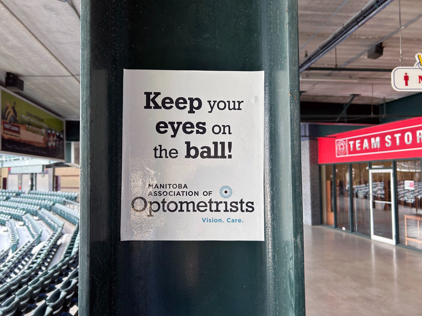 Sign on a post at the baseball stadium says Keep your eyes on the ball, sponsored by Mantioba Assoc of Optometrists.